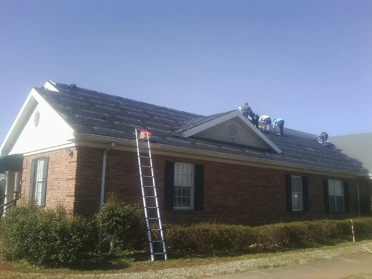 Crew tearing shingles off a brick ranch roof with a ladder set — Metal Roof Repair work by our crew in Kalona
