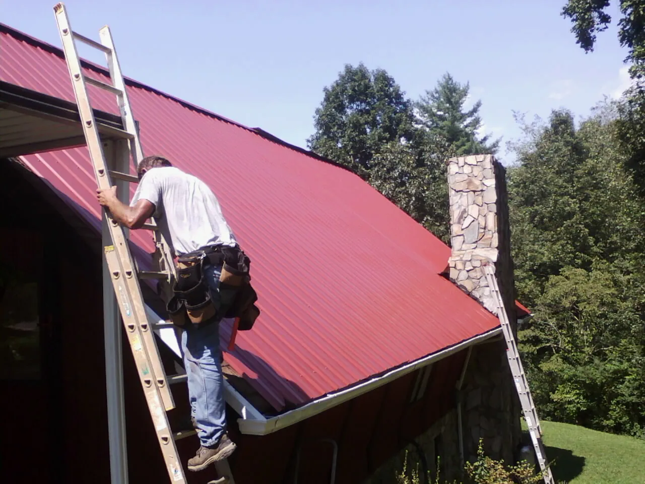 Roofer on a ladder beside a red metal roof and stone chimney — Soffit & Fascia Repair by Roofing Brothers in Kalona